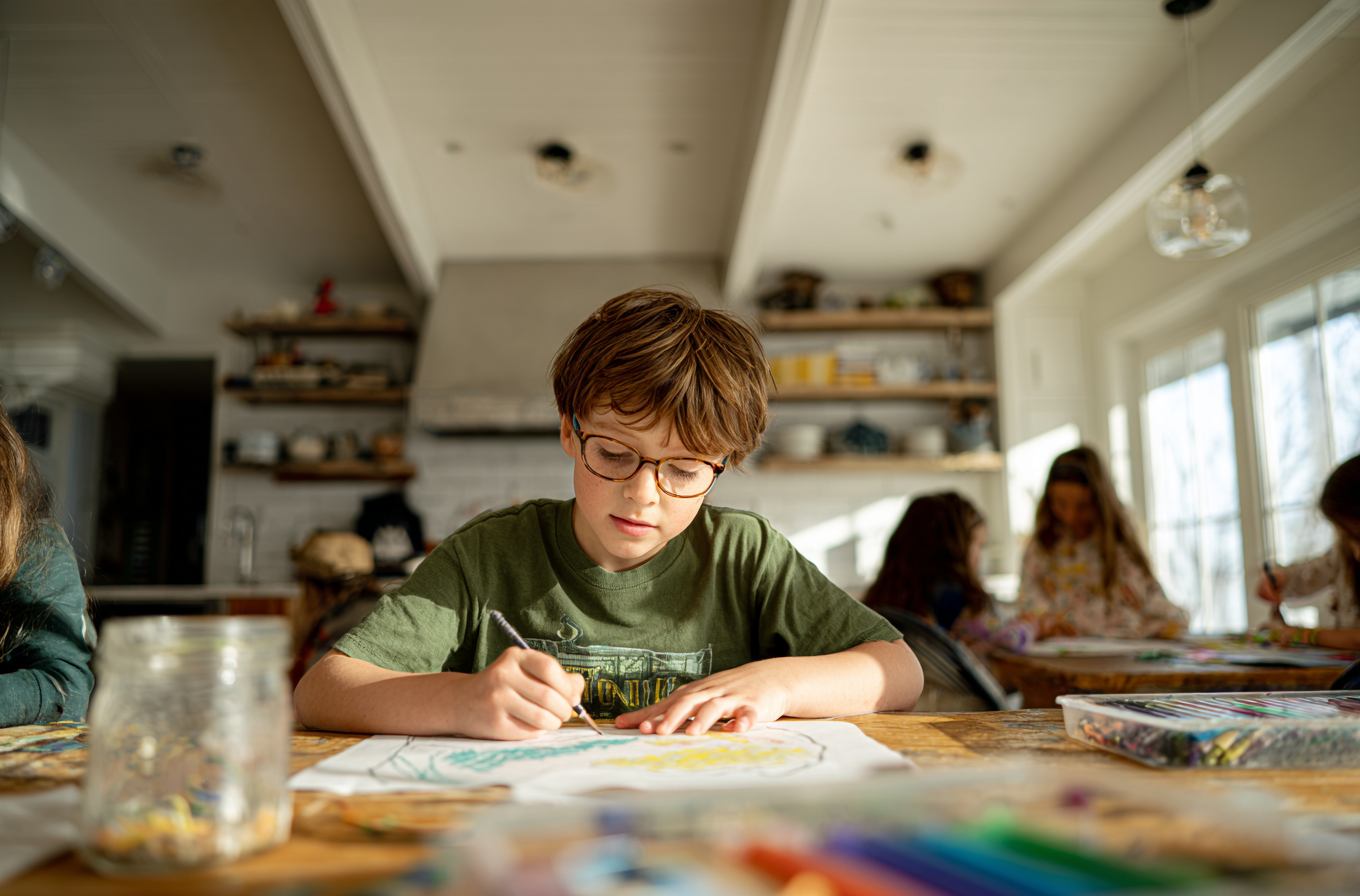 A student drawing at a kitchen table while other children work nearby